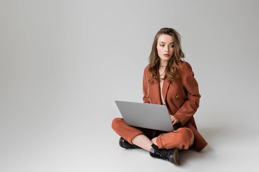 brunette young woman in golden necklace sitting with crossed legs in terracotta and trendy suit, using laptop while working remotely on grey background, freelancer, digital nomad, looking away - Photo, Image