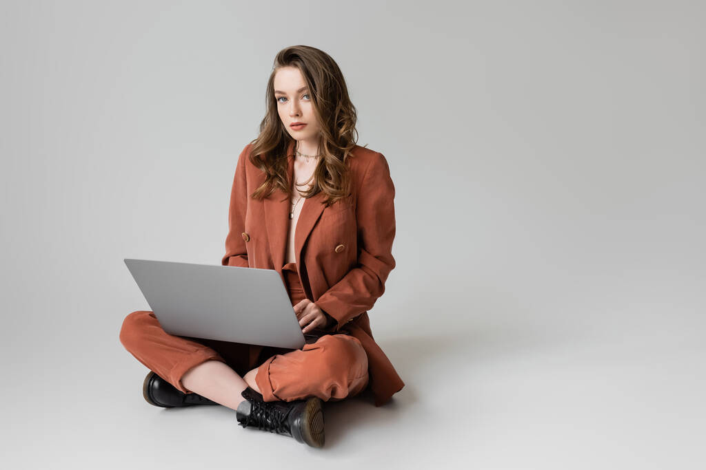 urocza młoda kobieta w złotym naszyjniku siedzi ze skrzyżowanymi nogami w terakocie i modnym garniturze, korzystając z laptopa podczas zdalnej pracy na szarym tle, freelancer, digital nomad  - Zdjęcie, obraz