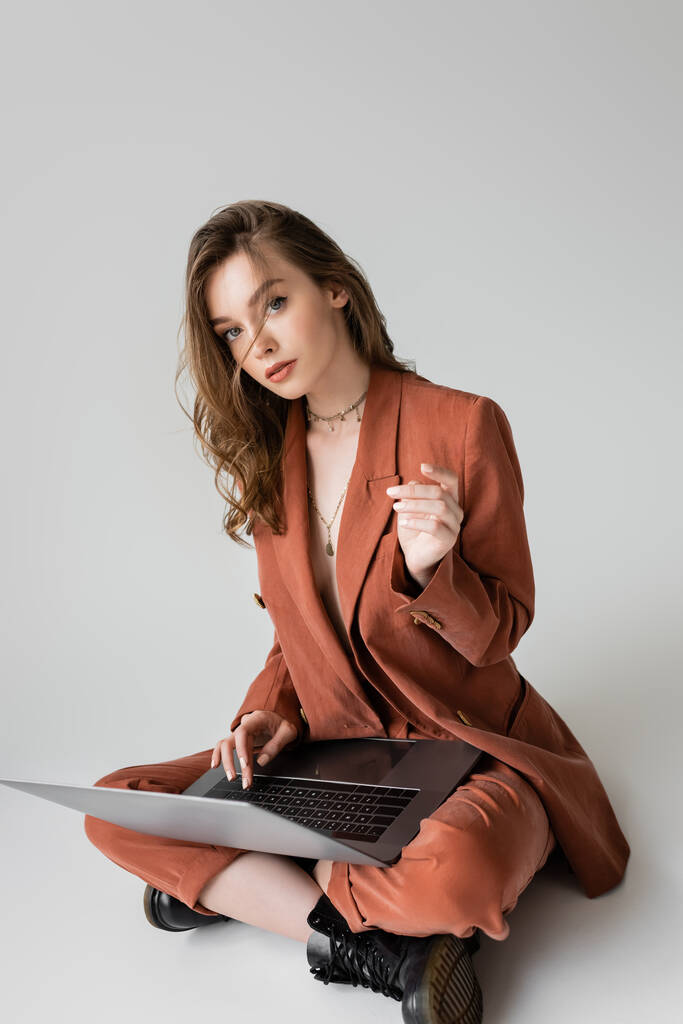 junge Frau in goldener Halskette sitzt mit überkreuzten Beinen im braunen und trendigen Anzug, benutzt Laptop, während sie ferngesteuert auf grauem Hintergrund arbeitet, Freiberuflerin, digitale Nomadin, schaut in die Kamera - Foto, Bild