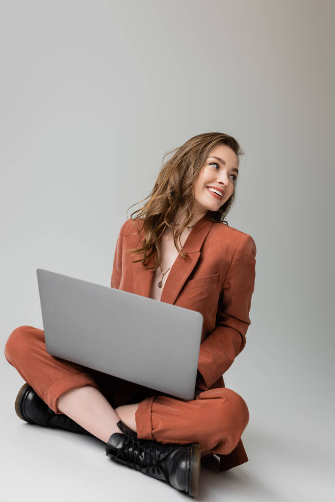 χαρούμενη νεαρή γυναίκα σε χρυσό κολιέ κάθεται με σταυρωμένα πόδια σε μοντέρνο κοστούμι, χρησιμοποιώντας το laptop, κοιτάζοντας μακριά, ενώ εργάζονται εξ αποστάσεως σε γκρι φόντο, ελεύθερος επαγγελματίας, ψηφιακή νομάδα  - Φωτογραφία, εικόνα