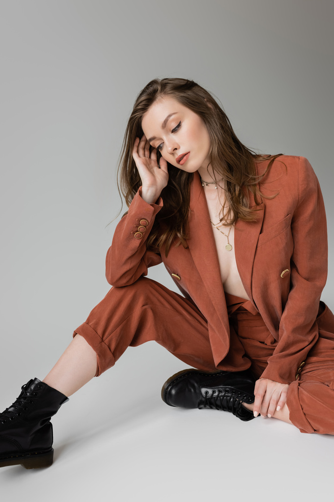 full length of sensual and young woman with brunette and wavy hair sitting in trendy and oversize suit with blazer, pants and black boots, looking down on grey background, stylish model   - Photo, Image