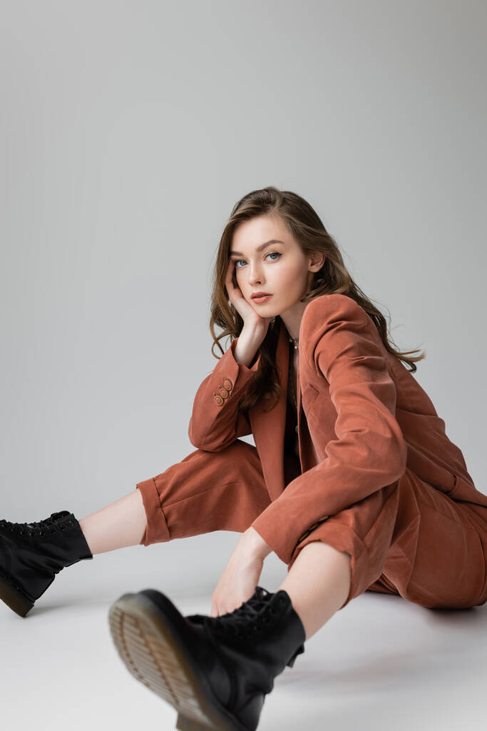 full length of fashionable model with brunette and wavy hair sitting in trendy and oversize suit with blazer, pants and black boots, looking at camera on grey background, stylish young woman  - Photo, Image