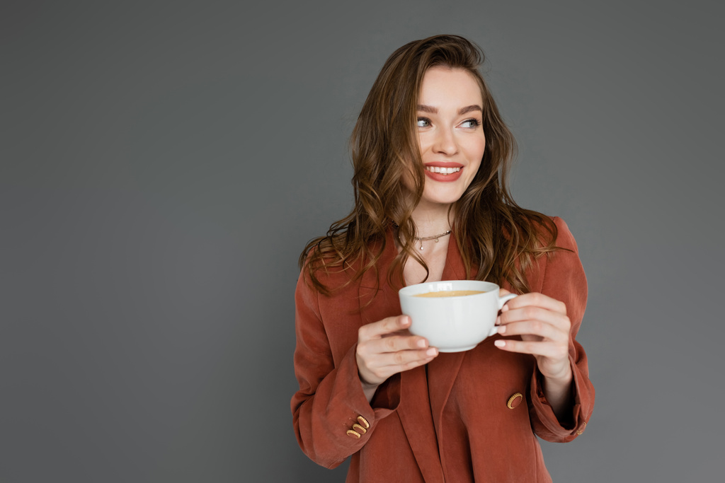 šťastná mladá žena s brunetkou vlasy na sobě hnědé a trendy oblek s blejzrem a drží šálek kávy při pohledu pryč na šedé pozadí, pracovní život rovnováhu - Fotografie, Obrázek