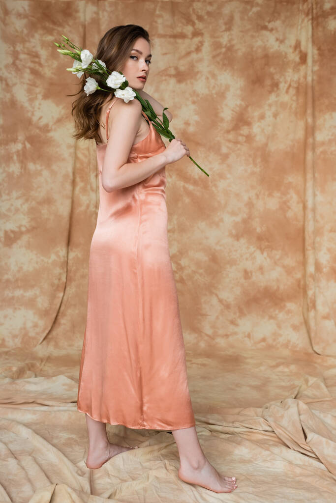 Çıplak ayaklı, esmer, pembe ipekli, beyaz çiçekli elbisesi olan genç bir kadın bej rengi arka planda, şehvetli, zarif ve sofistike duruyor.  - Fotoğraf, Görsel