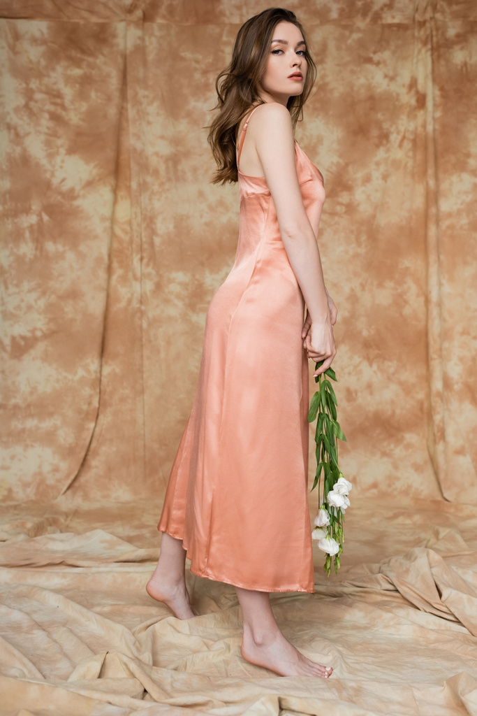 longitud completa de descalzo y morena y mujer joven en vestido de seda rosa deslizamiento sosteniendo flores blancas mientras mira a la cámara mientras está de pie sobre fondo beige moteado, sensualidad, elegancia  - Foto, imagen