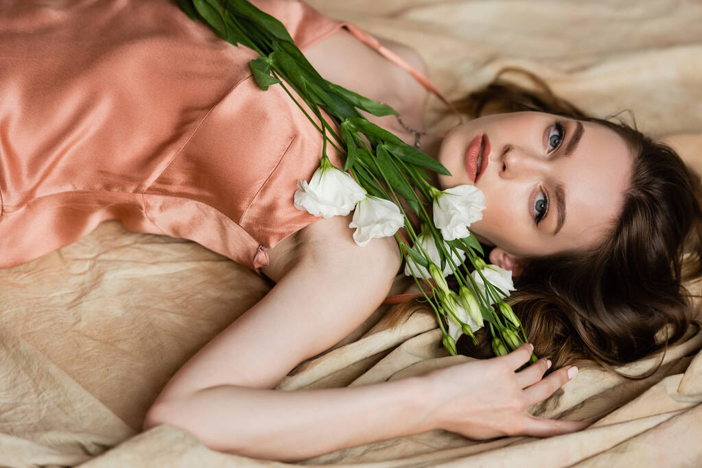 young woman in pink silk slip dress lying on linen fabric and holding white flowers on beige background, sensuality, sophistication, elegance, eustoma, looking at camera, graceful pose  - Photo, Image
