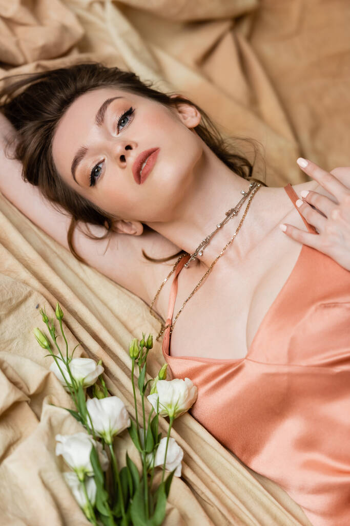 uwodzicielska młoda kobieta z brunetką leżąca w seksownej spódnicy na lnianej tkaninie w pobliżu delikatnych białych kwiatów i patrząca w kamerę, beżowe tło, wyrafinowanie, zmysłowość, elegancja, eustoma  - Zdjęcie, obraz