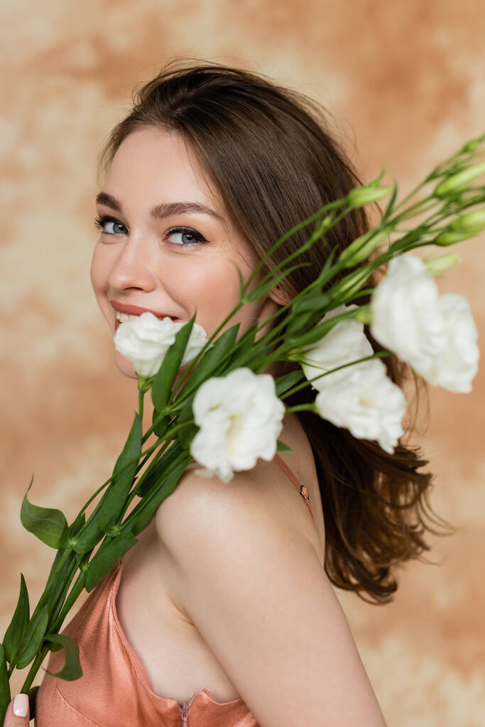 joyeuse jeune femme aux cheveux bruns posant en robe rose glissante et tenant des fleurs d'eustomie blanches sur fond beige marbré, sensualité, sophistication, élégance, regardant la caméra - Photo, image