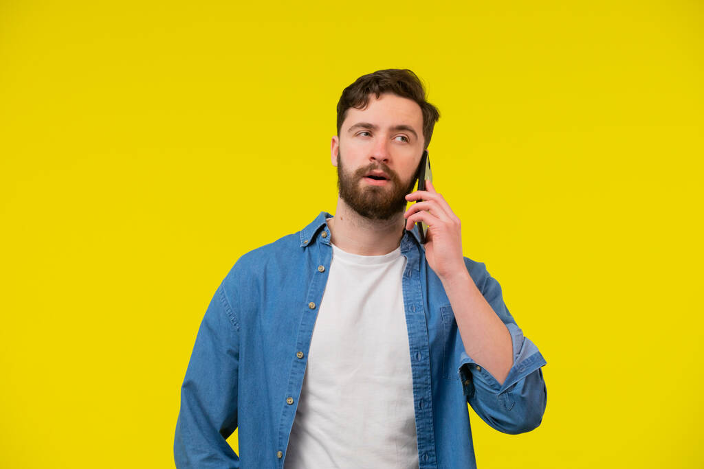 młody uśmiechnięty szczęśliwy wesoły biały mężczyzna 20s nosić biały casual koszula rozmowa mówić na telefon komórkowy prowadzenie przyjemny rozmowa patrzeć na bok odizolowany na zwykły żółty tło studio portret. - Zdjęcie, obraz