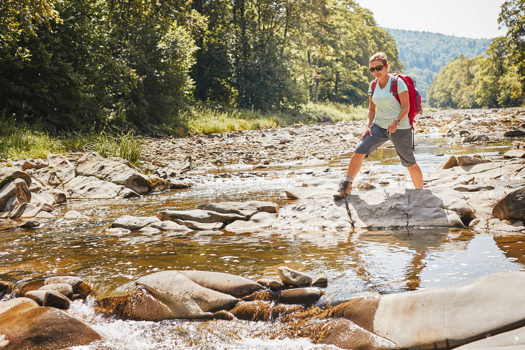 Πεζοπορία με την εικόνα έννοια σακίδιο. Γυναίκα backpacker φορώντας μπότες πεζοπορία διασχίζοντας το βουνό ποτάμι. Γυναίκα πεζοπορία στα βουνά κατά τη διάρκεια του καλοκαιρινού ταξιδιού. Ταξίδι διακοπών κοντά στη φύση. Φυσικό τοπίο - Φωτογραφία, εικόνα