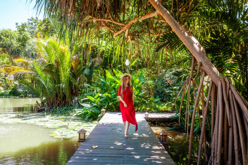 Reizende vrouw in rode jurk ontspannen in tropische resort. Vrouwelijke reiziger op exotische zomervakantie. Meisje met strohoed lopend op houten brug nabij meer in tuin met palmbomen en groene planten - Foto, afbeelding