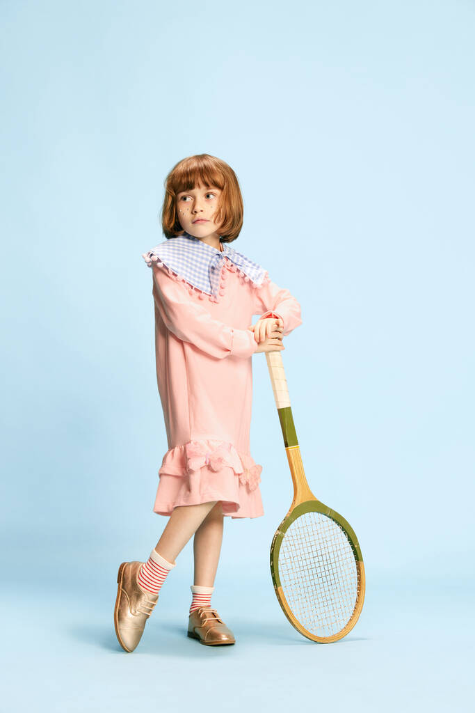 Full-length portret van mooi klein meisje in roze jurk poseren met tennis racket tegen blauwe studio achtergrond. Concept van kindertijd, emoties, plezier, mode, actieve levensstijl, sport - Foto, afbeelding