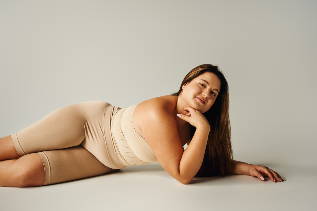 smiling woman in strapless top with bare shoulders and underwear posing while lying in studio on grey background, body positive, self-love, plus size, figure type, looking at camera  - Photo, Image