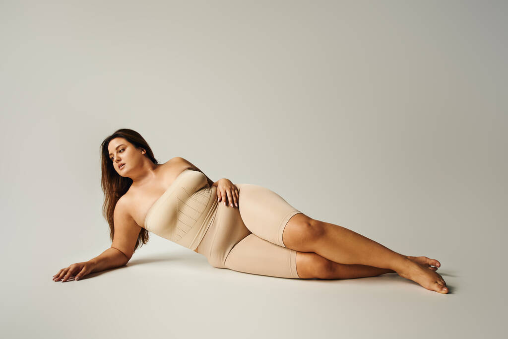 bosá žena s plus velikosti těla v bezramenném svršku s holými rameny a spodní prádlo pózuje při ležení ve studiu na šedém pozadí, tělo pozitivní, self-love, relaxační, dívá se jinam  - Fotografie, Obrázek