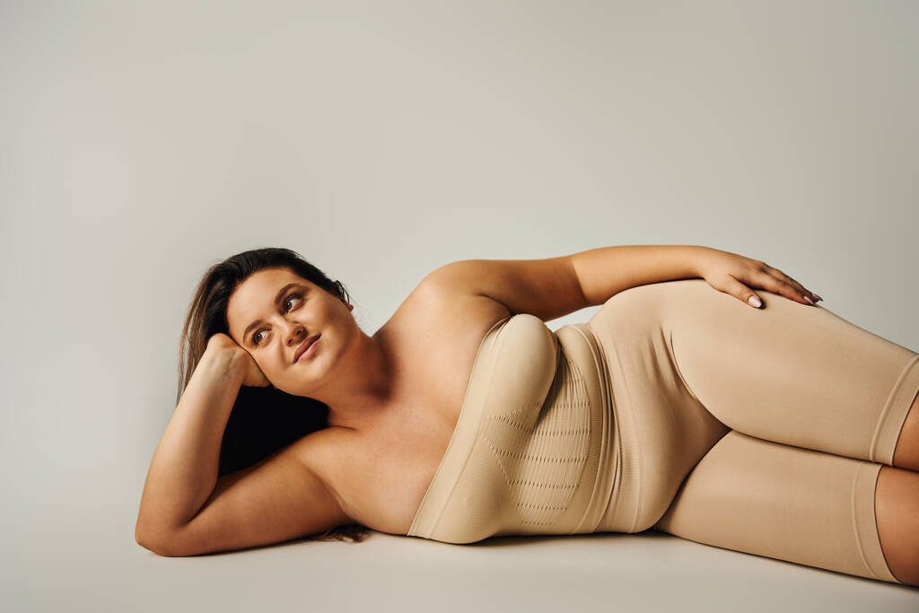 brünette Frau mit Plus-Size-Körper in trägerlosem Top mit nackten Schultern und Unterwäsche posiert im Studio auf grauem Hintergrund, körperbetont, selbstverliebt, entspannend, wegschauend  - Foto, Bild