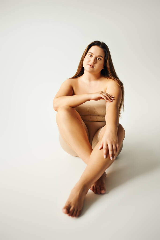 Barfuß-Model mit Plus-Size-Körper in trägerlosem Top mit nackten Schultern und Unterwäsche posiert im Studio auf grauem Hintergrund, körperbetont, selbstverliebt, entspannend, in die Kamera schauend  - Foto, Bild