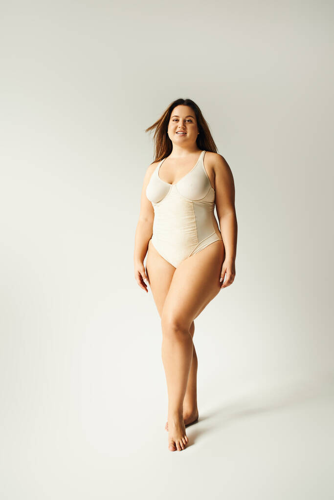 pełna długość boso i plus size kobieta w beżowym body suit pozowanie podczas stania w studio na szarym tle, body positive, typ figury, poczucie własnej wartości, uśmiech patrząc w obiektyw  - Zdjęcie, obraz