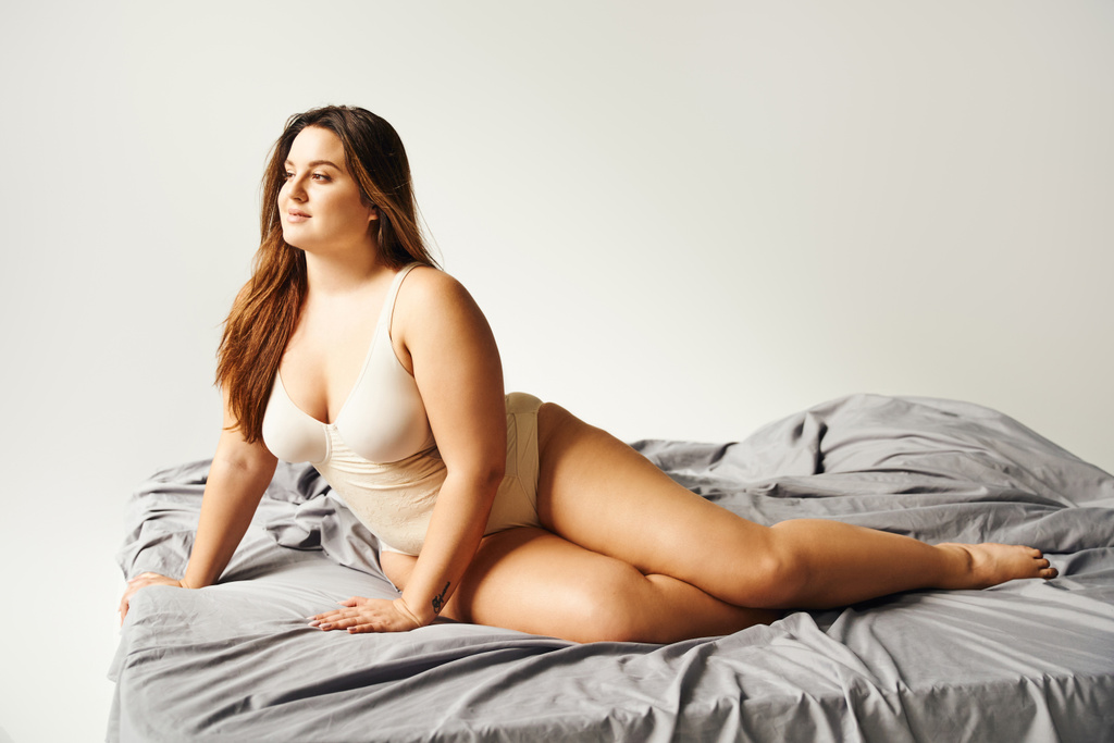 dreamy brunette plus size woman with natural makeup wearing beige bodysuit and posing on bed with grey bedding while looking away, body positive, figure type, tattoo translation: harmony  - Photo, Image