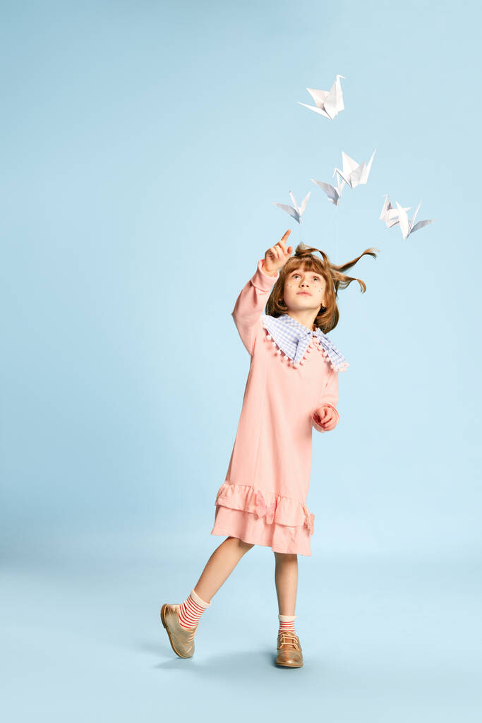 Ganztägiges Porträt eines verspielten, hübschen kleinen Mädchens in rosa Kleid, das mit Papiervögeln vor blauem Studiohintergrund spielt. Konzept der Kindheit, Emotionen, Spaß, Mode, Lifestyle, Fantasie - Foto, Bild