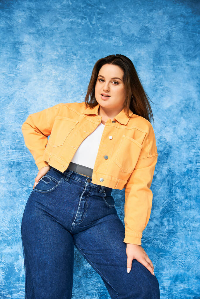 woman with plus size body and long hair, wearing crop top, orange jacket and denim jeans while posing with hand on hip and looking at camera on mottled blue background, body positive  - Photo, Image