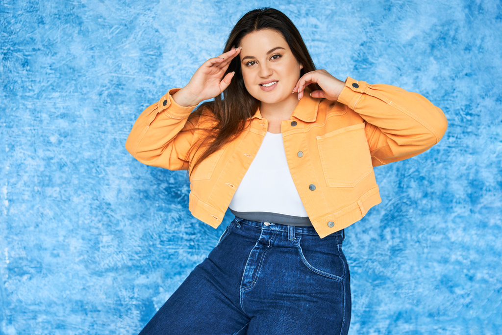 Self Acceptance, body positive, happy plus size woman with long hair and natural makeup wearing crop top, orange jacket and denim jeans while posing and looking at camera on mottled blue background  - Photo, Image