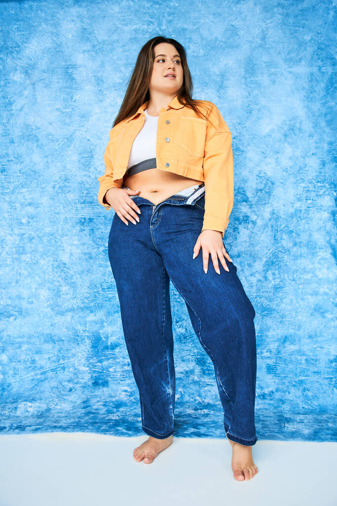 full length of barefoot body positive woman with plus size body and brunette hair posing in orange jacket, crop top and denim jeans while posing and looking at camera on mottled blue background  - Photo, Image