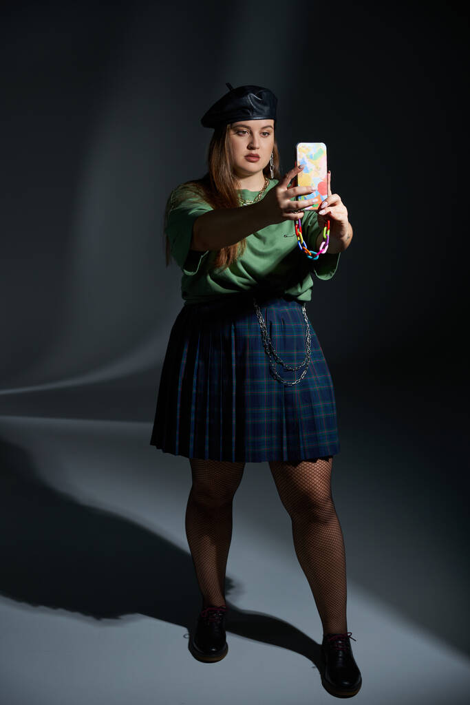 plus size woman posing in leather beret, green t-shirt, plaid skirt with chains, fishnet tights and black shoes, taking selfie on smartphone on dark background, tattoo translation: harmony - Photo, Image