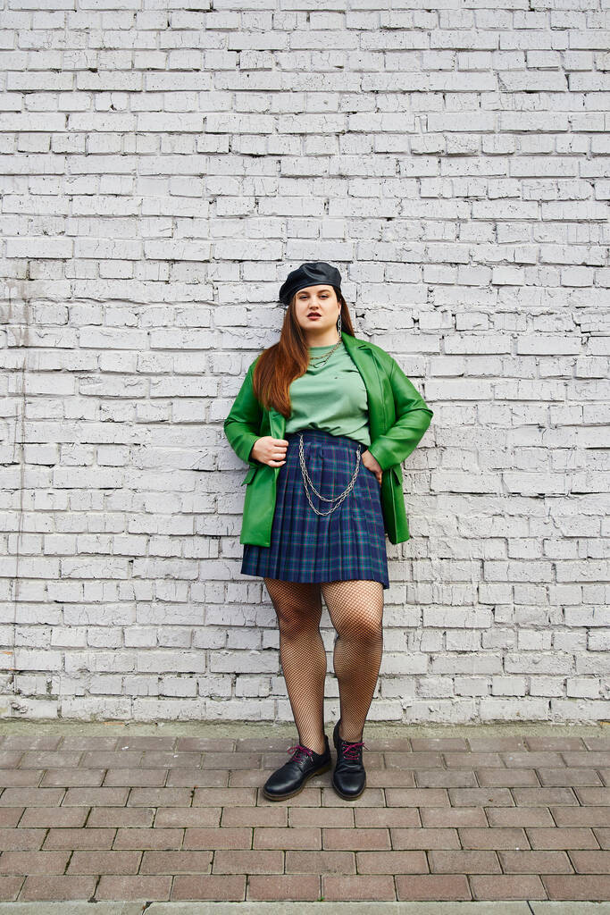 full length of brunette plus size woman posing in leather jacket with beret, plaid skirt with chains, fishnet tights and black shoes while standing near brick wall on urban street  - Photo, Image
