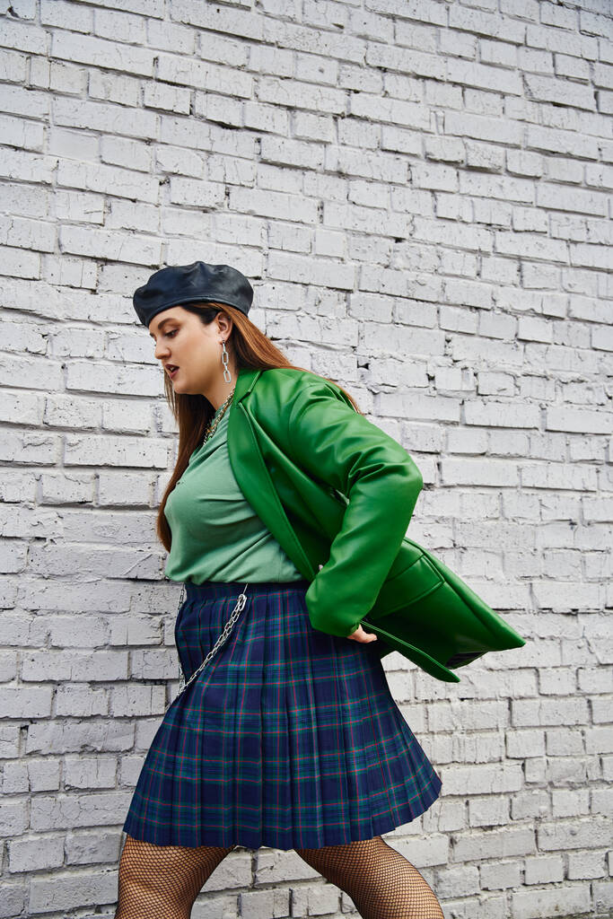 zijaanzicht van stijlvolle plus size vrouw in groene leren jas, zwarte baret, geruite rok met kettingen en visnet panty 's lopen in de buurt van baksteen muur op stedelijke straat, lichaam positief, zelf-liefde, stedelijke chique - Foto, afbeelding