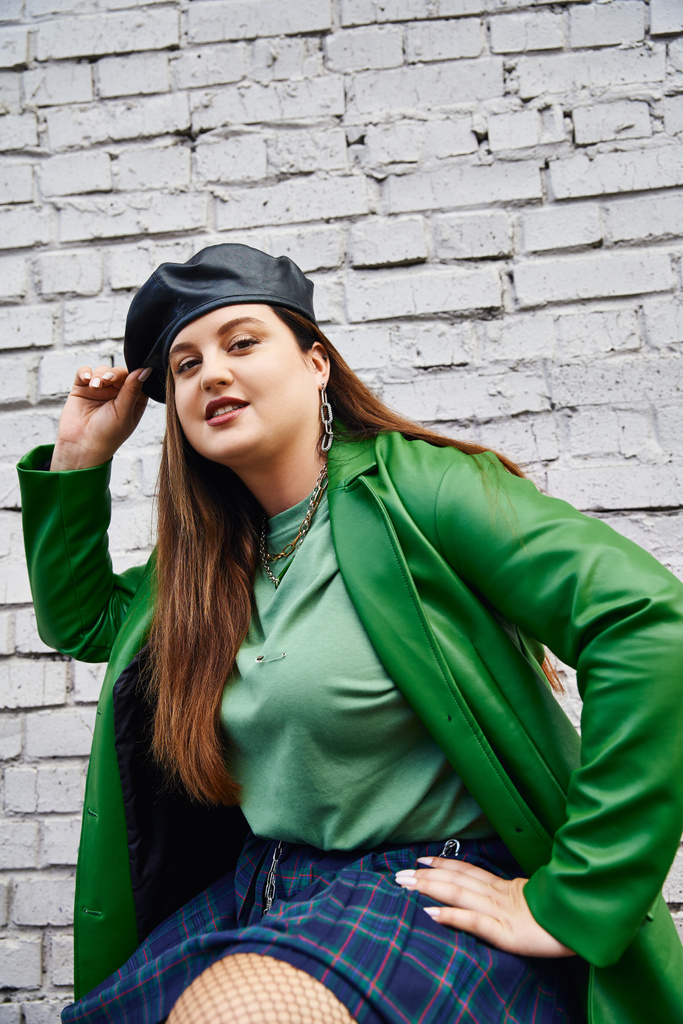 mujer elegante más tamaño en chaqueta de cuero verde sonriendo mientras toca boina negra y posando en falda a cuadros y medias de malla cerca de la pared de ladrillo en la calle urbana, cuerpo positivo, amor propio, urbano elegante - Foto, imagen