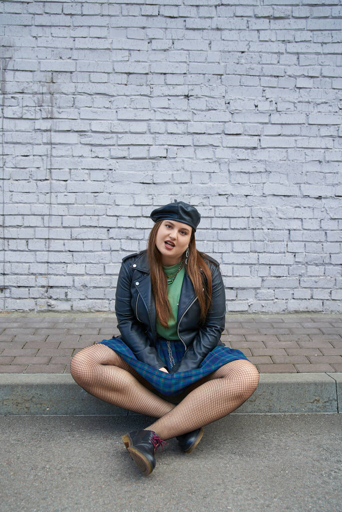 full length of chic plus size woman posing in leather jacket, plaid skirt with chains, fishnet tights and black shoes while sitting near brick wall on urban street, body positive, full length - Photo, Image