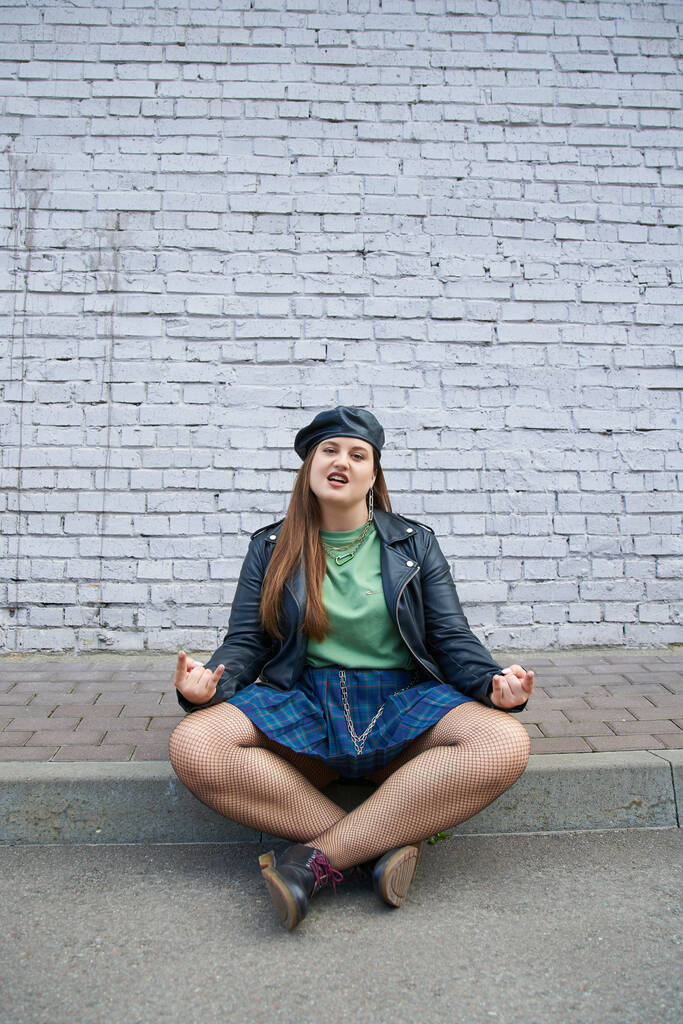 chic plus size woman posing in leather beret and jacket, plaid skirt with chains, fishnet tights and black shoes while sitting with crossed legs near brick wall on urban street, rock sign - Photo, Image
