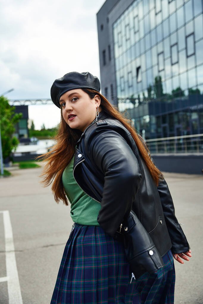stylish woman with plus size body walking in leather jacket with black beret, plaid skirt and greet t-shirt near blurred modern building on urban street outdoors, body positive, looking at camera - Photo, Image