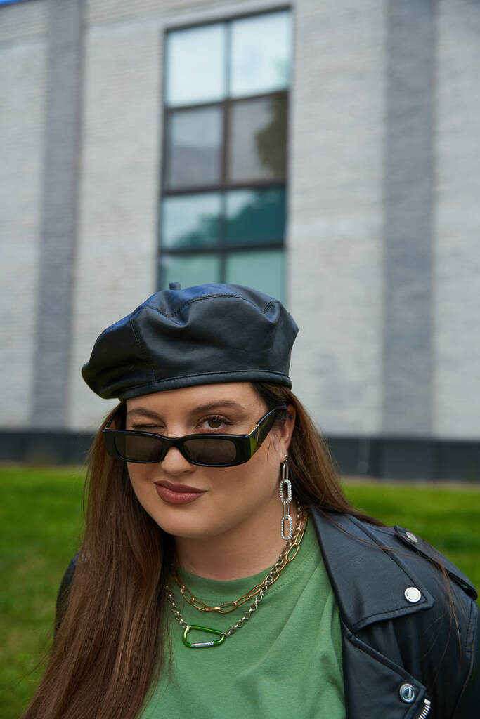 portrét plus velikost brunetky žena pózující ve stylových slunečních brýlích, kožená bunda s černým baretem a pozdrav tričko mrkání v blízkosti rozmazané moderní budovy na městské ulici venku, tělo pozitivní  - Fotografie, Obrázek