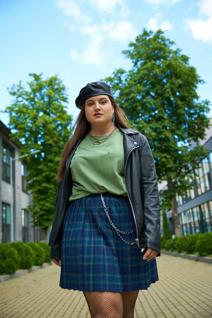 low angle view of confident woman with plus size body posing in leather jacket with black beret near blurred modern buildings and alley with trees on urban street outdoors, body positive  - Photo, Image