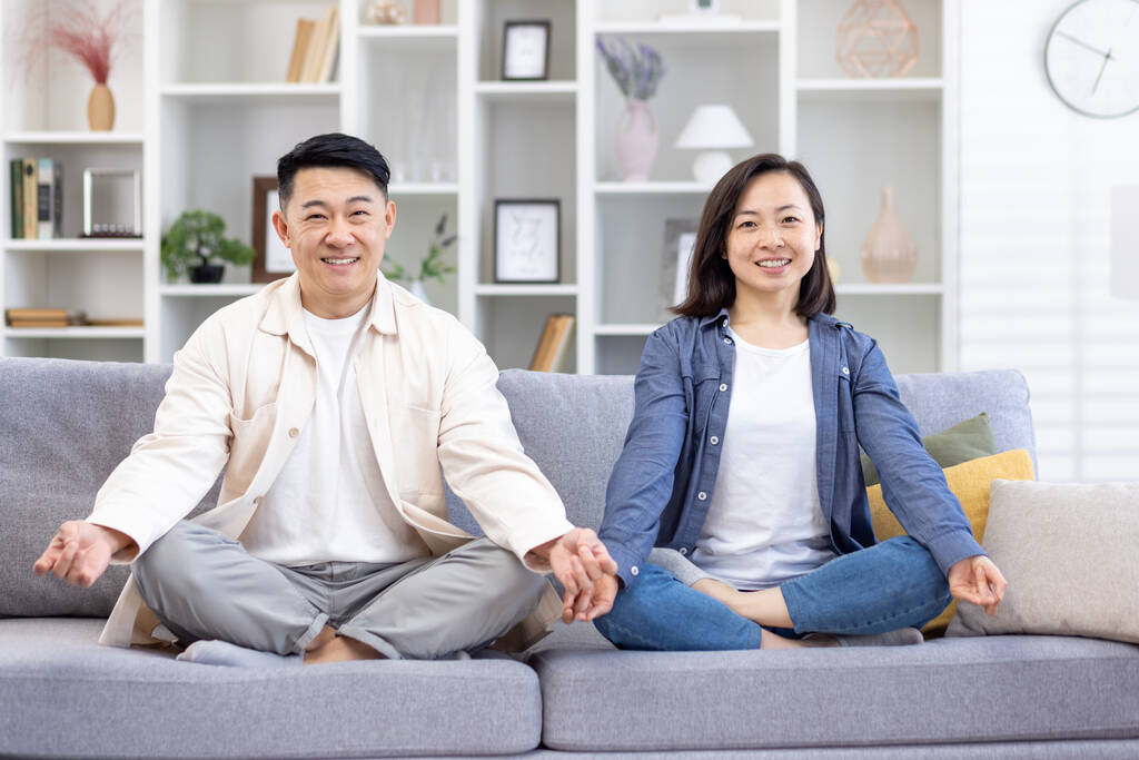 Портрет молодой азиатской семьи, пара, занимающаяся йогой дома. Мужчина и женщина сидят на диване в позе лотоса и улыбаются в камеру. заботиться об их психическом здоровье. - Фото, изображение