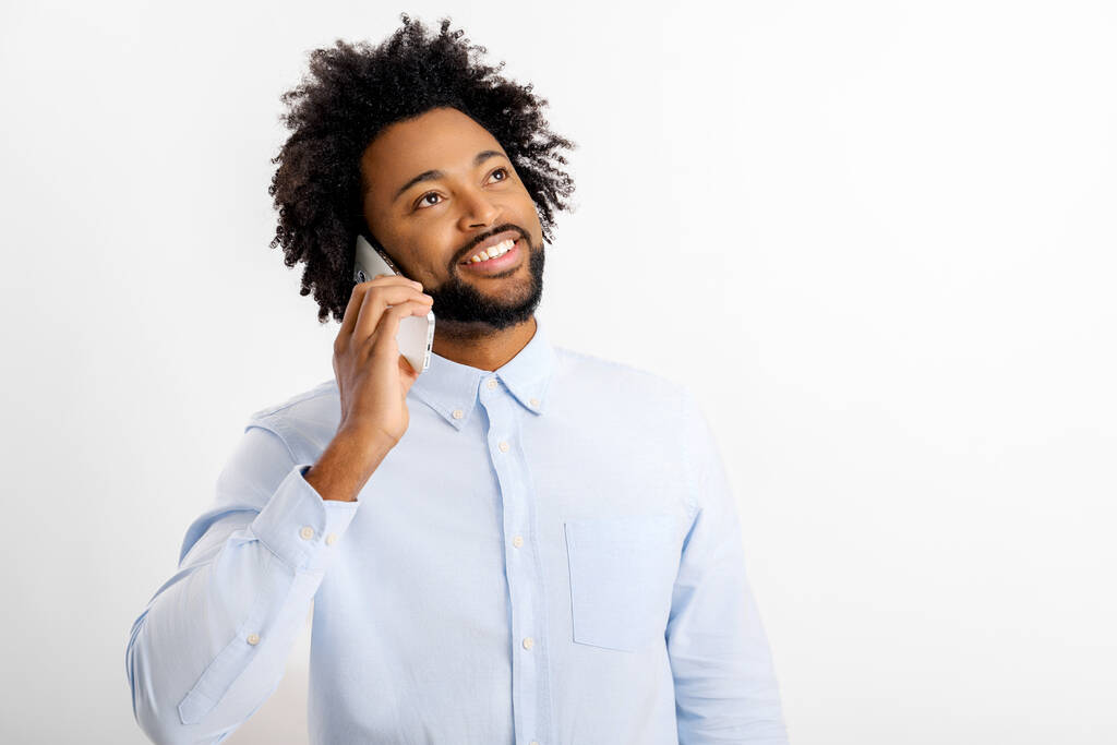 Kıvırcık, güler yüzlü beyaz gömlekli mutlu Afro-Amerikan adam cep telefonuyla konuşuyor, beyaz arka planda izole edilmiş hoş bir sohbet yürütüyor, stüdyo portresi. - Fotoğraf, Görsel