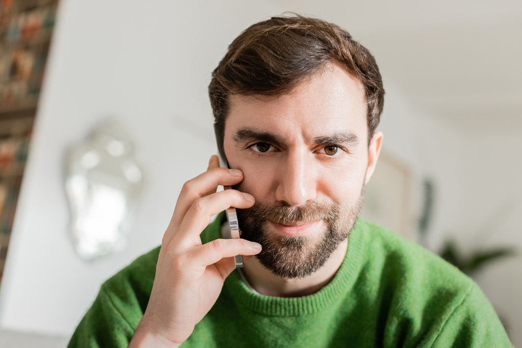 Portrait d'homme brune et barbu en pull vert parlant sur smartphone et regardant la caméra - Photo, image