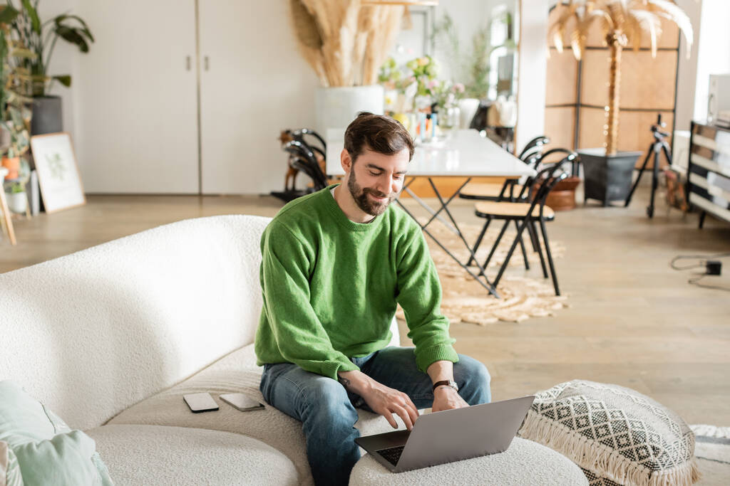 Freelancer barbudo alegre en jersey y jeans trabajando en el portátil mientras está sentado cerca de los teléfonos inteligentes - Foto, imagen