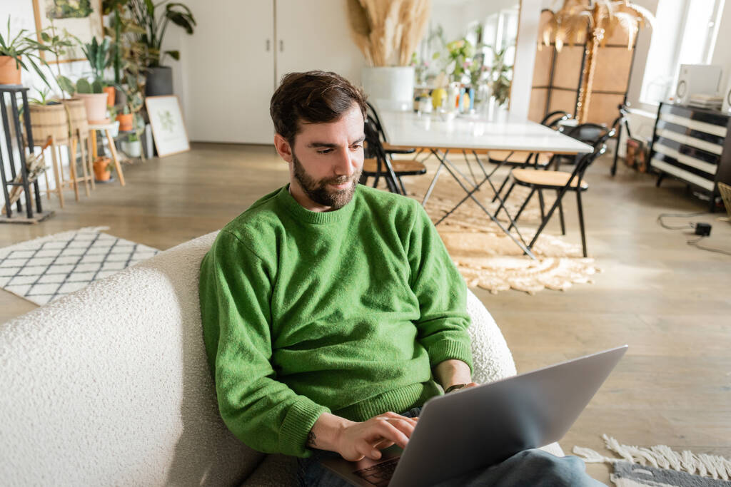 baard en brunette freelancer in groen jumper met behulp van laptop tijdens het werken in huis  - Foto, afbeelding
