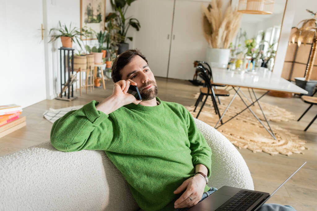 Γενειοφόρος και μελαχρινή freelancer σε πράσινο πουλόβερ και τζιν μιλάμε για smartphone στο σαλόνι - Φωτογραφία, εικόνα