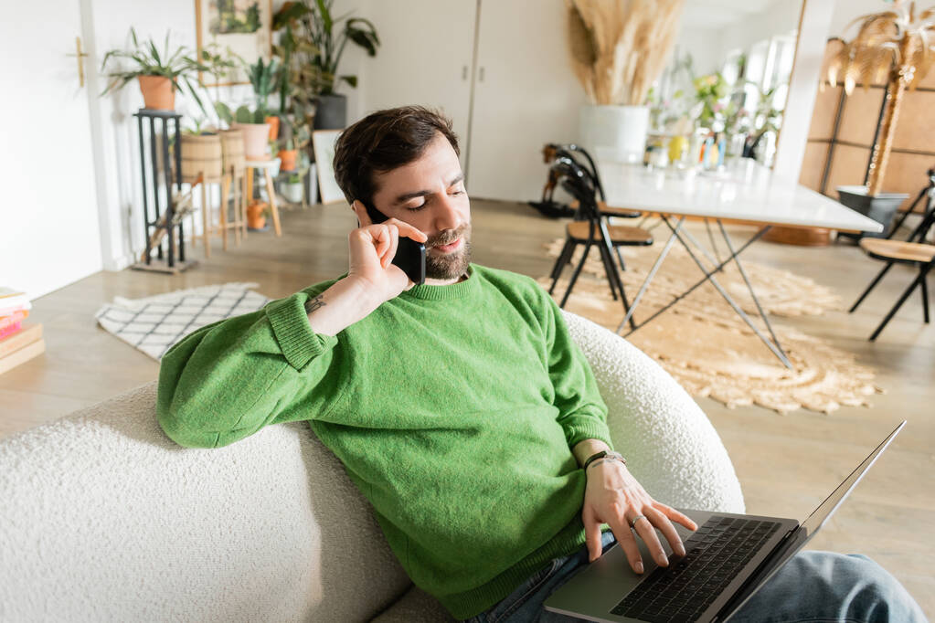 Γενειοφόρος ελεύθερος επαγγελματίας σε πράσινο jumper και τζιν μιλώντας στο smartphone, ενώ χρησιμοποιώντας φορητό υπολογιστή στο σπίτι  - Φωτογραφία, εικόνα