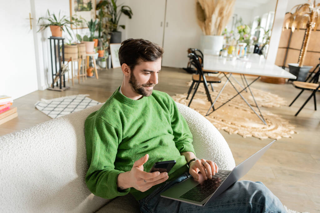 Smiling pigiste barbu en pull vert et jeans à l'aide d'un ordinateur portable et tenant smartphone à la maison  - Photo, image