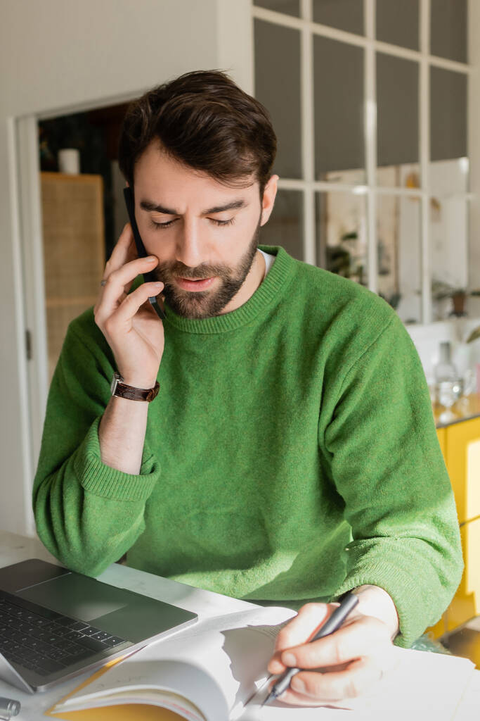 Portré szakállas vállalkozó zöld jumper beszél okostelefon és írás notebook  - Fotó, kép