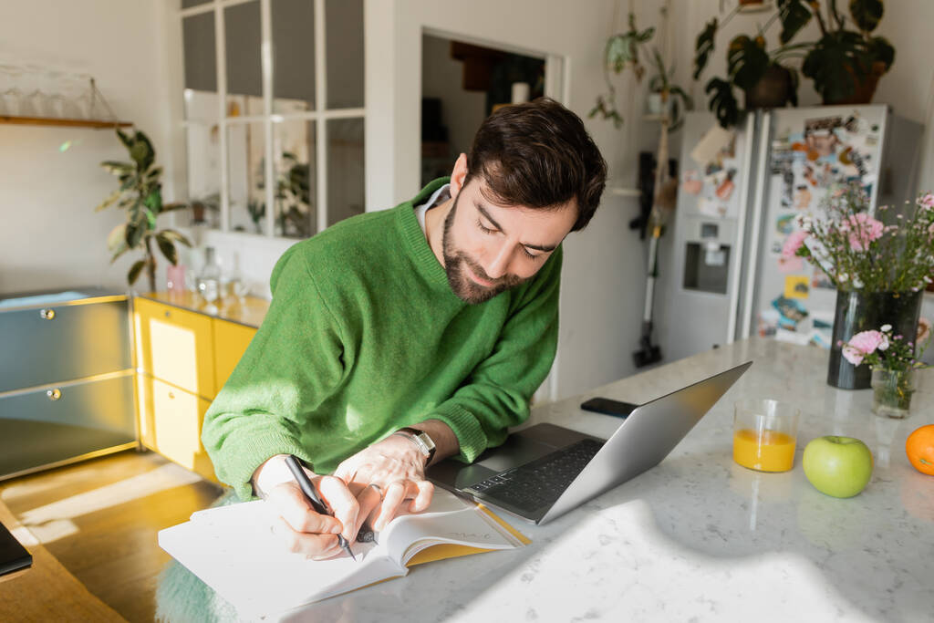 Bearded freelancer in green jumper writing on notebook while working near devices at home  - Photo, Image