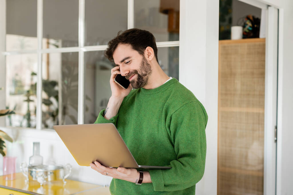 Позитивная брюнетка и бородатый фрилансер в свитере разговаривают на смартфоне и держат ноутбук дома  - Фото, изображение