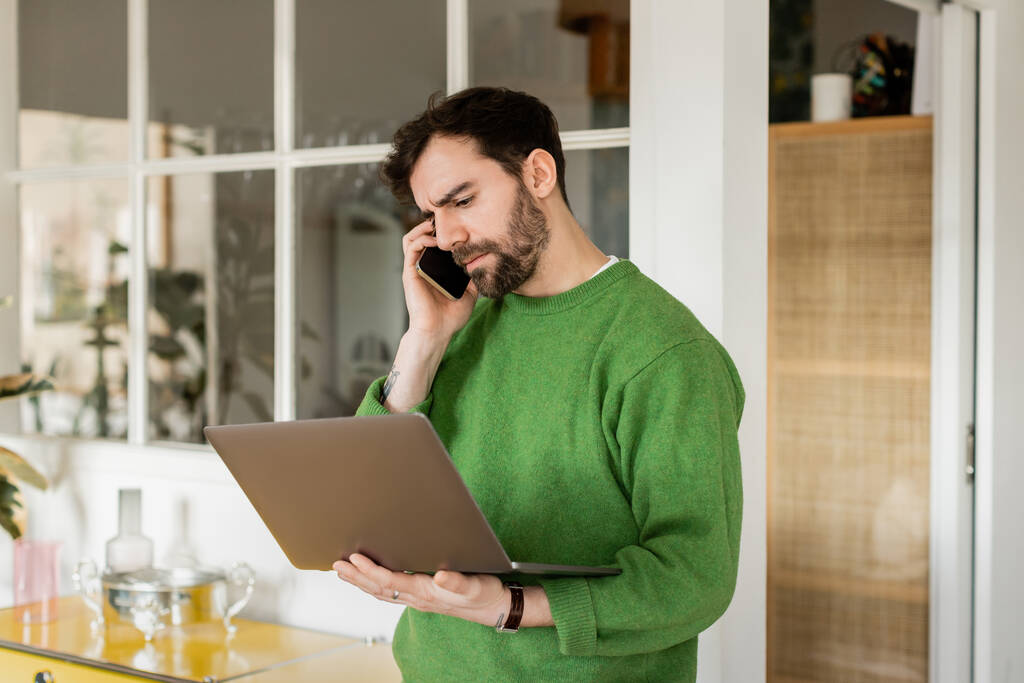 Freelancer enfocado en jumper verde que sostiene el portátil y habla en el teléfono inteligente en casa  - Foto, Imagen