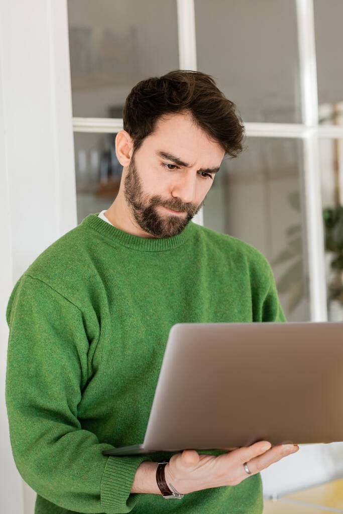 Portré fókuszált és szakállas szabadúszó alkalmi zöld jumper nézi laptop, távoli munka - Fotó, kép