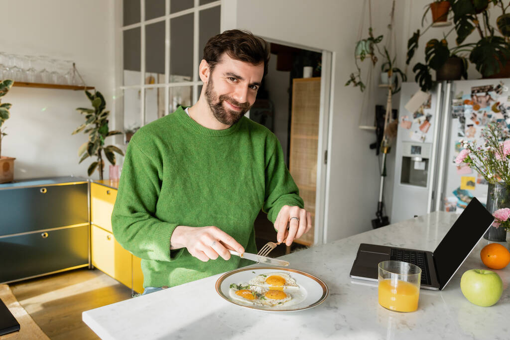 Yeşil kazaklı neşeli sakallı adam elinde çatal bıçak takımıyla modern mutfakta kahvaltı hazırlıyor.  - Fotoğraf, Görsel