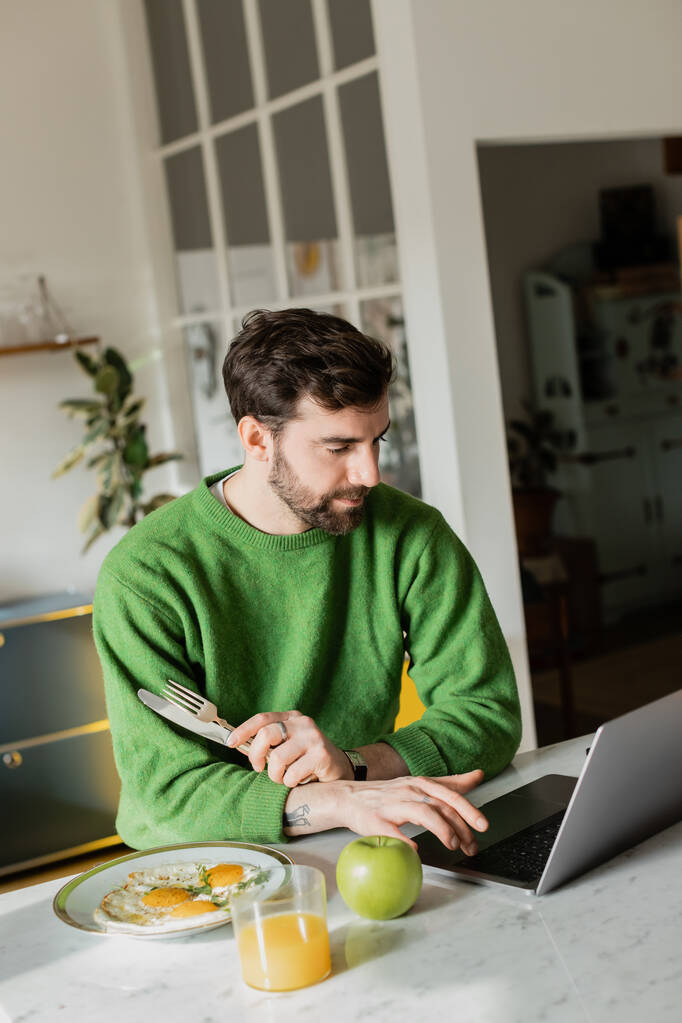 Baard en getatoeëerde freelancer in trui met bestek en laptop thuis  - Foto, afbeelding
